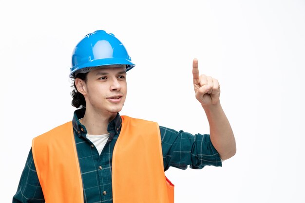 Pleased young male engineer wearing safety helmet and safety vest looking at side pointing finger up isolated on white background