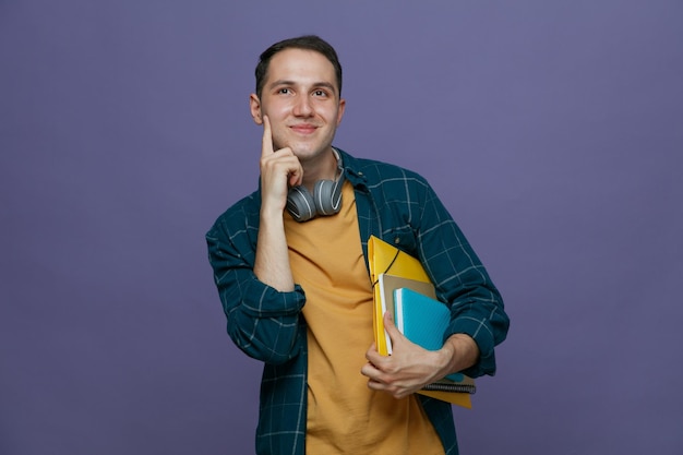 Pleased young male student wearing headphones around neck holding study tools under arm keeping hand on chin looking up isolated on purple background