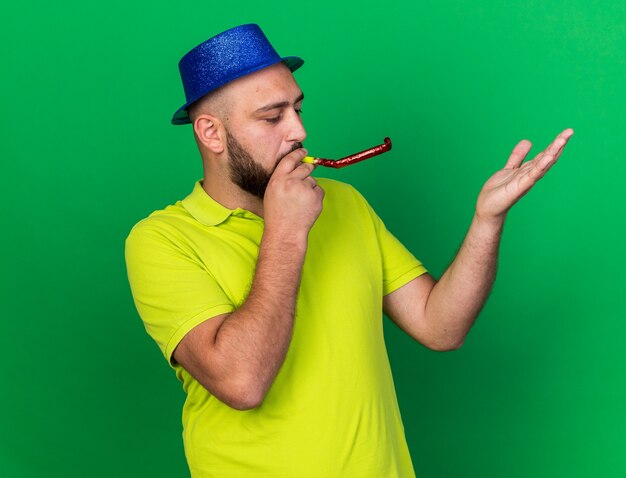 Pleased young man wearing blue party hat blowing party whistle raising hand isolated on green wall