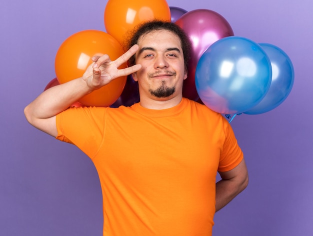 Pleased young man wearing orange t-shirt standing in front balloons showing peace gesture isolated on purple wall