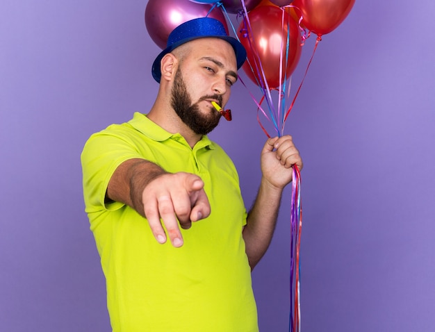 Pleased young man wearing party hat holding balloons blowing party whistle points at camera 