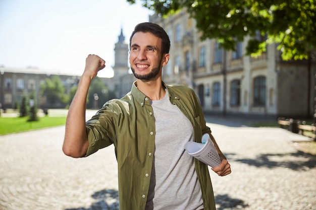 Довольный молодой человек с тетрадью в руке стоит перед зданием университета