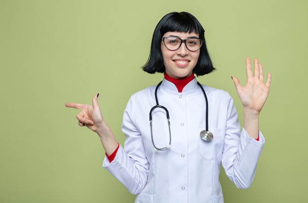 Pleased young pretty caucasian girl with optical glasses in doctor uniform with stethoscope pointing at side and keeping hand open 