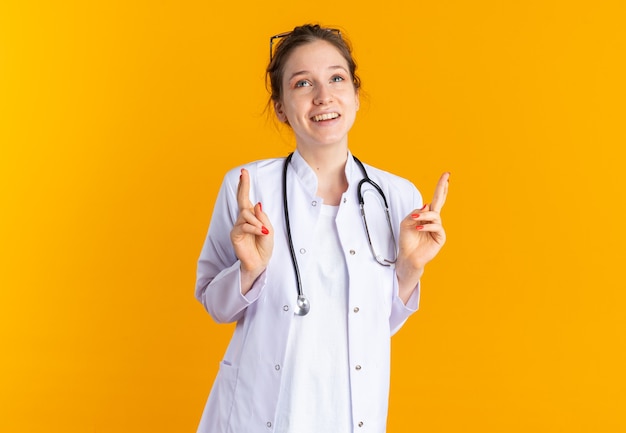 Pleased young slavic girl in doctor uniform with stethoscope crossing fingers looking up isolated on orange wall with copy space