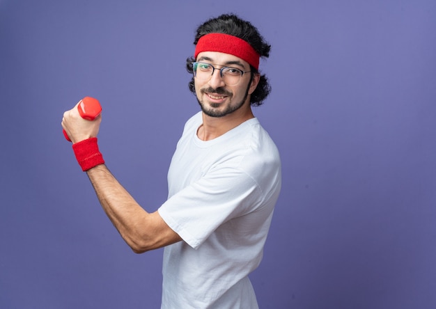 Pleased young sporty man wearing headband with wristband exercising with dumbbells 