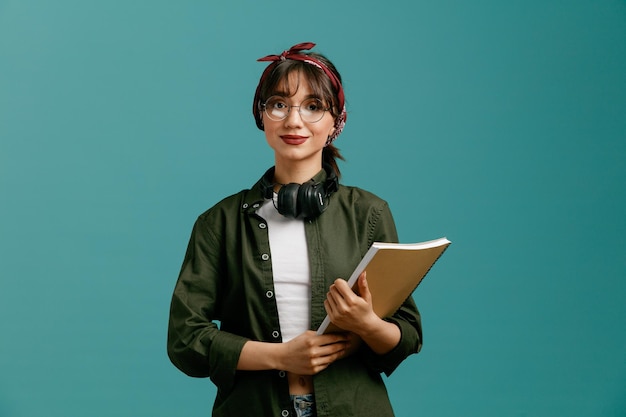 Pleased young student girl wearing bandana glasses and headphones around neck holding large note pad with both hands looking at camera isolated on blue background