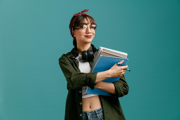 Pleased young student girl wearing bandana glasses and headphones around neck holding large note pads and pen looking at camera isolated on blue background