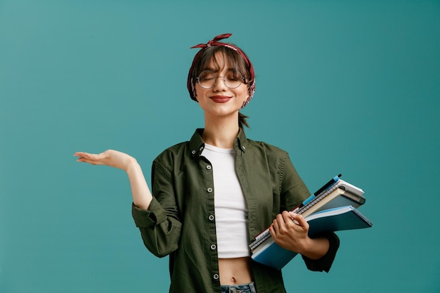 Photo pleased young student girl wearing bandana glasses holding note pads with pen showing empty hand with closed eyes isolated on blue background