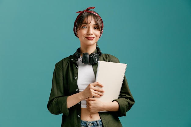 Pleased young student girl wearing bandana sunglasses and headphones around neck holding large note pad with both hands looking at camera isolated on blue background