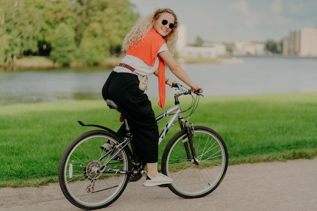 Pleased young woman enjoys new route on bicycle, rides among\
lake, green lawn and buildings far away into distance, wears summer\
shades, casual outfit, white sneakers, being in good physical\
shape