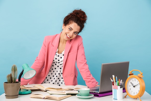 Photo pleased young woman smiling and looking at camera while working at desk isolated over blue wall