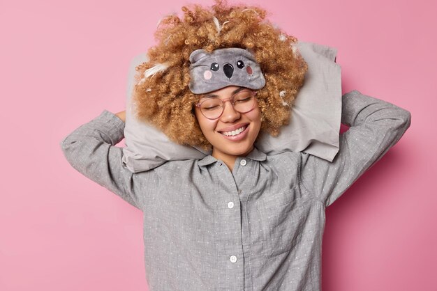 Pleased young woman with pleasant smile on face keeps hands\
behind head leans on soft pillow dressed in nightwear sees sweet\
dreams takes nap isolated over pink background people and rest\
concept