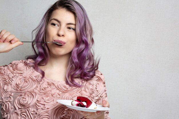 Pleased young woman with purple hair eating delicious cheese cake over grey background. Space for text