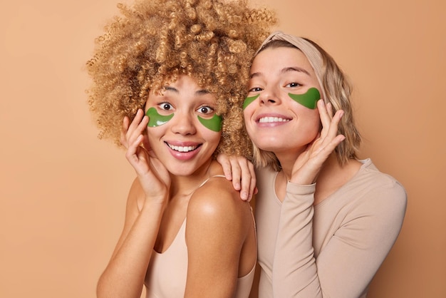 喜んでいる若い女性は、肌に栄養を与えるために緑色のヒドロゲルパッチを適用し、若くて美しく見えたいと思っています。茶色の背景に対して互いに密接に立っているパーティーの準備をします。美容マスク