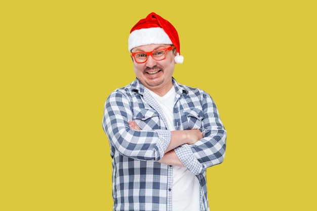 Pleasure happy modern middle aged man in red santa cap, eyeglasses and in checkered shirt standing with crossed arms and toothy smiling, looking at camera. Indoor, isolated on yellow background.