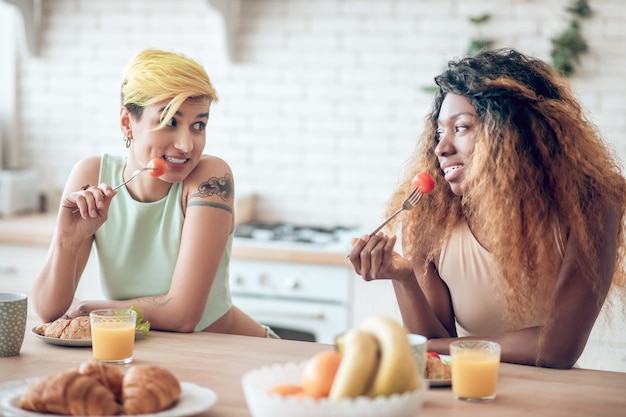 Plezier samen. twee vrolijke jonge vriendinnen in vrijetijdskleding in speelse bui aan het ontbijt met vorken met tomaten