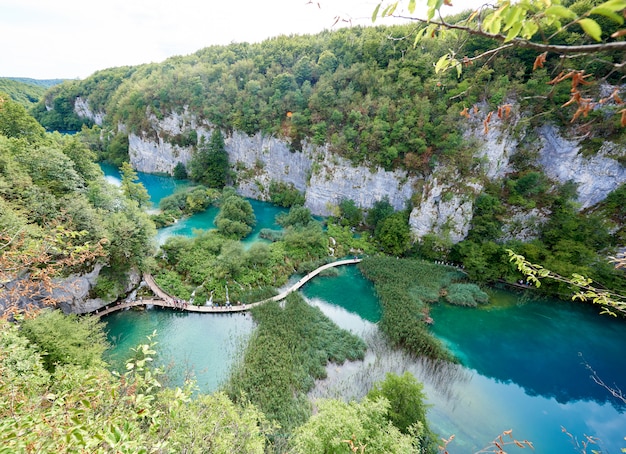 Plitvice Lakes National Park - UNESCO World Heritage Centre. Aerial view of lit by sun green forest on rocky cliffs and long wooden bridge with tourists over lakes with clear blue-green water.