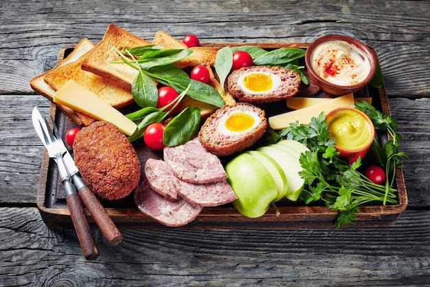 Photo ploughman's lunch of toasts, cheddar cheese wedges, apple, scotch eggs, sliced headcheese, tomatoes, spinach, hot mustard served on a wooden tray on a rustic table, horizontal view from above