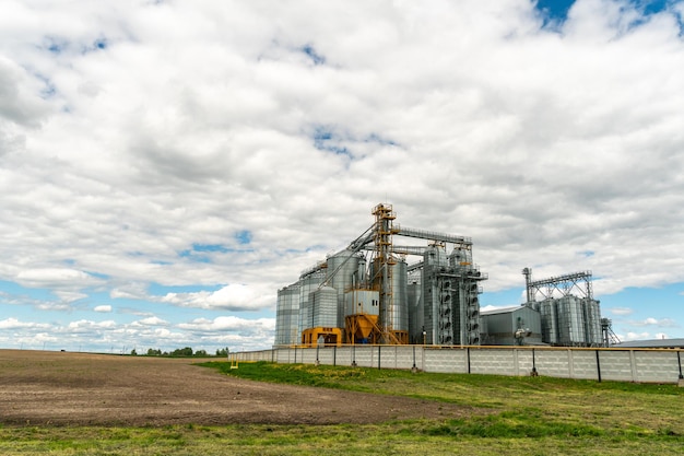 Next to the plowed agricultural field installed silver silos on\
agro manufacturing plant for processing drying cleaning and storage\
of agricultural products flour cereals and grain granary\
elevator