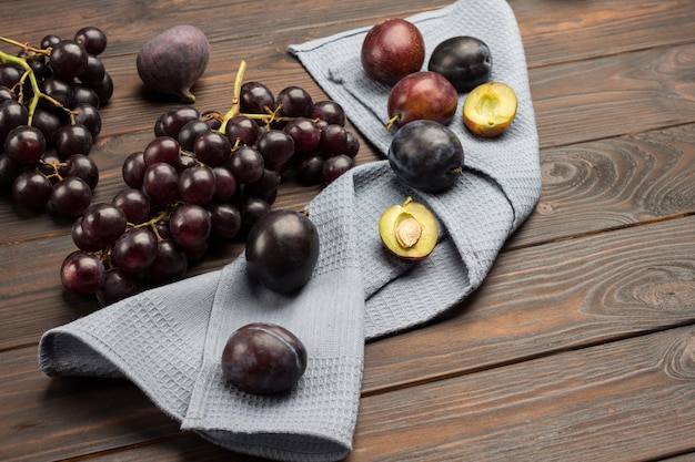 Plum fruits on gray napkin. Half plum with pit. Bunches of black grapes on table. Dark wooden surface. Top view. Copy space