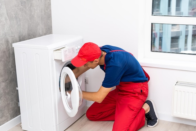 Plumber in overalls with tools is repairing a washing machine in the house