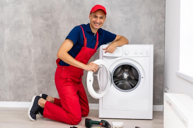 Plumber in overalls with tools is repairing a washing machine in the house