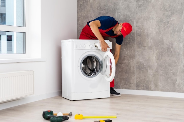 Plumber in overalls with tools is repairing a washing machine\
in the house