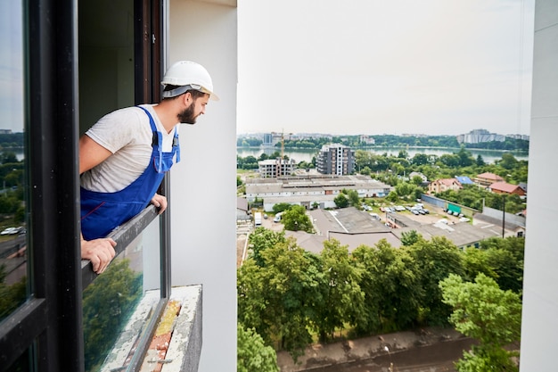 青いオーバーオールと白いヘルメットを着て窓の外を眺める配管工 新しい建物のアパートでの設置作業中に新鮮な空気を得る コピー スペース