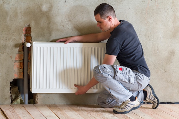 Plumber worker installing heating radiator in an empty room of a newly built apartment or house.