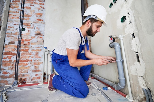 Plumber working with grey sewer pipes fixing them to wall with a help of screwdriver Side view of man standing on knees wearing blue uniform and white helmet Plumbing works home renovation concept