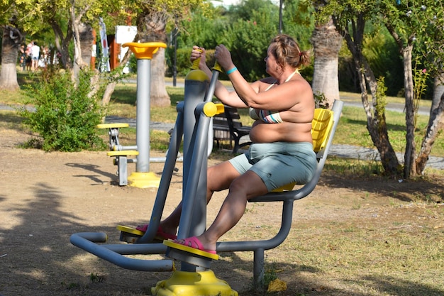 Plump fat woman in age doing exercises on simulators outside. fat woman pensioner doing sports on the street