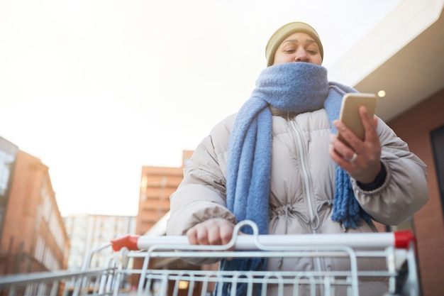 Plump woman going shopping with cart and reading a message on her mobile phone outdoors