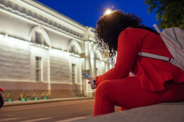 A plump woman in a red bright suit with a phone in her hands sits in the center of a European city