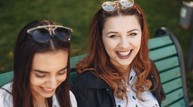 Plump woman with red hair looking up to camera and smile while sitting on the bench near her brunette friend with eyeglasses and relaxing