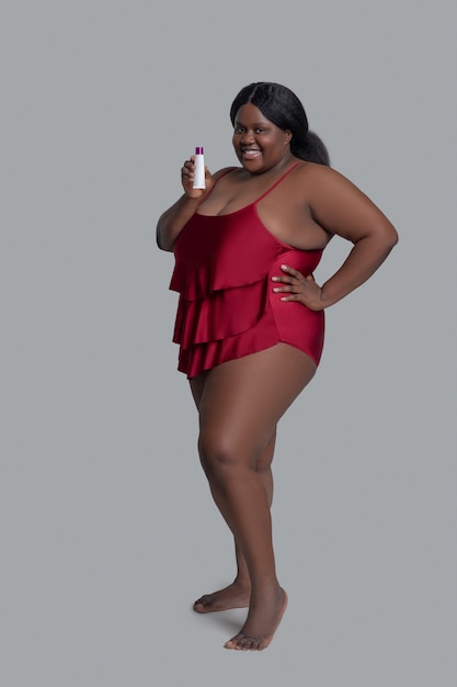 Plus size giovane donna afro-americana in rosso che tiene un tubo con crema e sorridente