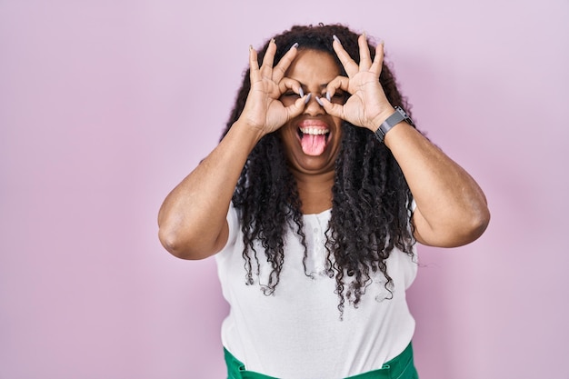 Plus size hispanic woman standing over pink background doing ok gesture like binoculars sticking tongue out eyes looking through fingers crazy expression