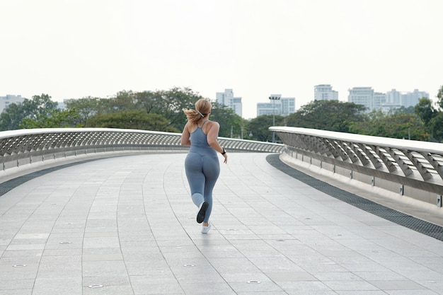 Plus size jonge vrouw joggen op lege brug vroeg in de ochtend, uitzicht vanaf de achterkant, vastberadenheid concept