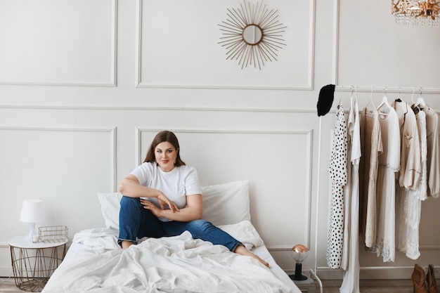 Plus-size model girl in jeans and white t-shirt sitting on the bed near hanger with clothes. Young smiling plump woman making a decision what to wear. XXL fashion