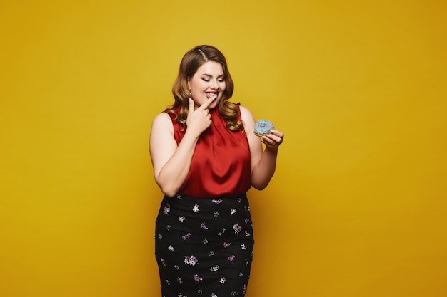 Plus-size model woman in a red satin blouse and black skirt ready to eat a tasty donut, isolated