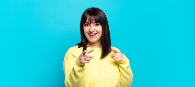 Plus size pretty woman smiling with a positive, successful, happy attitude pointing to the camera, making gun sign with hands