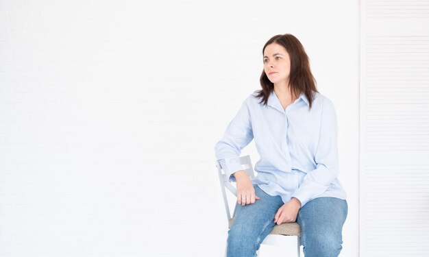 Plus size woman in blue shirt and jeans posing in studio on chair, white background with copy space.