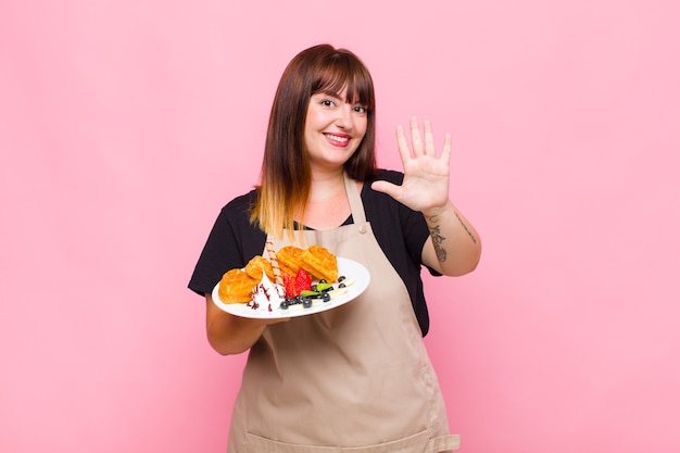 Plus size woman smiling and looking friendly, showing number five or fifth with hand forward, counting down