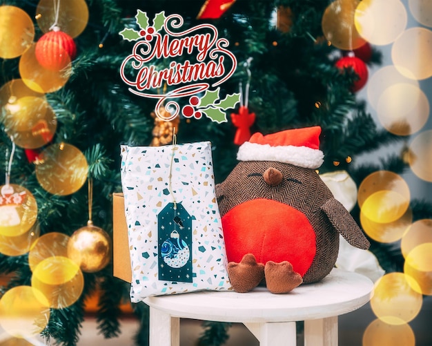 Plush robin toy with a Santa hat on a white stool near the Christmas tree decorated with ornaments