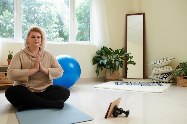 Plussize jonge vrouw volgt les op tabletcomputer bij het beoefenen van yoga thuis