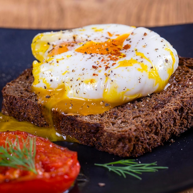 Poached egg on a piece of bread with fried red tomatoes on a wooden table close up