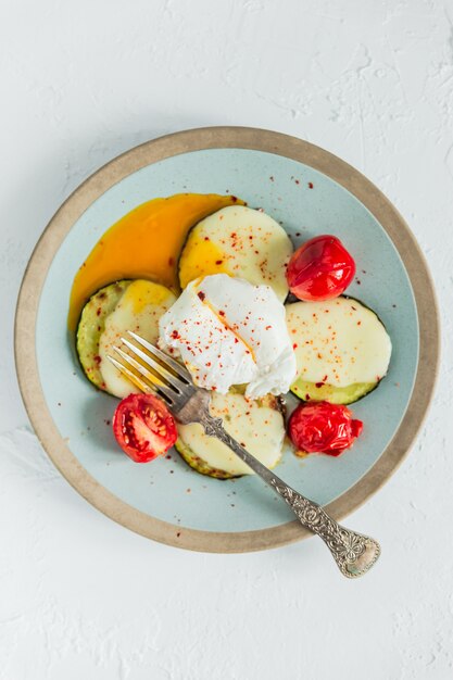 Poached egg with fried zucchini, mozzarella cheese, tomatos and red chili flakes, running yolk, fork. Top view.