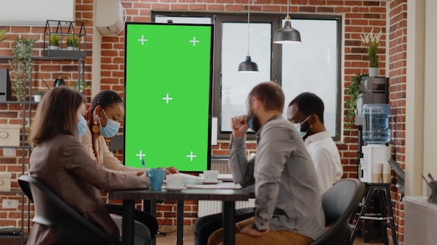 フェイスマスクを持った人々が、ビジネスオフィスで緑色の画面のモニターを見て、孤立したモックアップテンプレートと、covid19パンデミックの間にコピースペースに空白のクロマキーの背景を使用しています。
