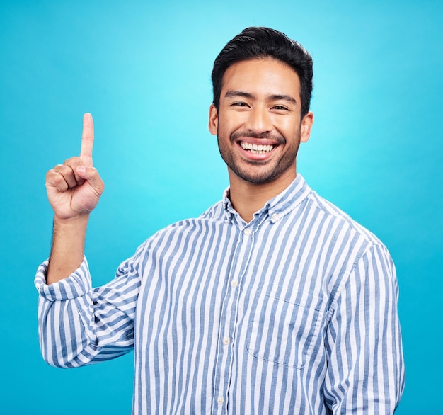 Point excited and portrait of Asian man on blue background for news information and announcement Advertising studio mockup and happy male pointing for copy space promotion and showing gesture