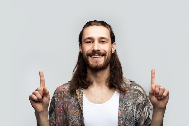 Point fingers up over his head with a smiling face to place text, logo or advertisement. Hipster traveler stylish carefree man on a white studio background, people lifestyle