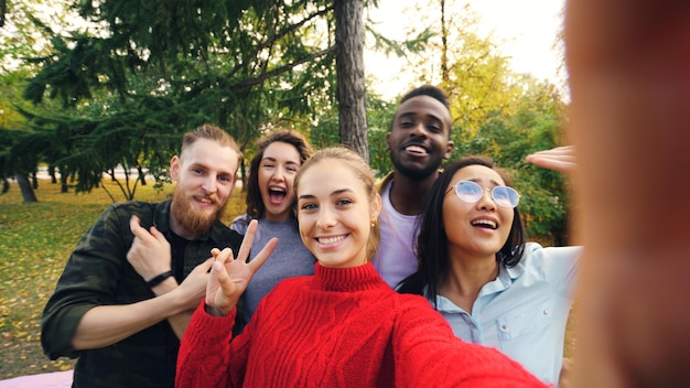 Photo point of view shot of young woman holding device with camera and taking selfie with friends multiethnic group in park in autumn photography and people concept